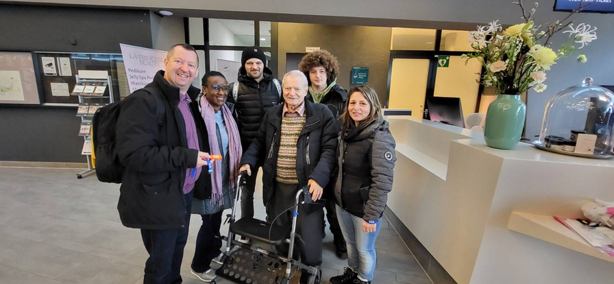 Sechs Personen posieren lächelnd in einer Lobby mit einem Rollator.