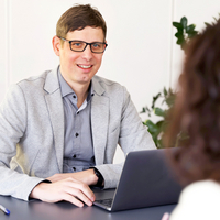 Eine lächelnde Person mit einem Laptop berät eine andere Person zum Thema 24-Stunden-Betreuung.