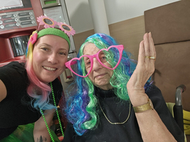 Frau mit bunten Perücken und Herzbrille lächelt gemeinsam mit älterer Dame.