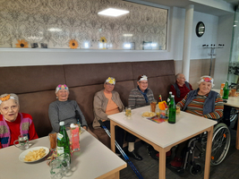 Senioren in einer gemütlichen Runde tragen bunte Papierhüte und genießen Snacks und Getränke.