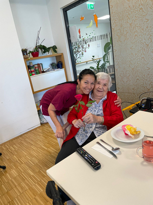 Eine lächelnde Frau umarmt eine ältere Dame mit einer Rose in der Hand.