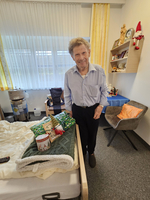 Ein älterer Herr steht in einem hellen Zimmer neben einem Bett mit Geschenken.