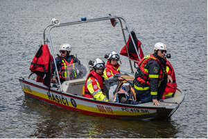 Ein Rettungsboot mit fünf Personen in Schutzkleidung fährt auf einem Gewässer. Die Einsatzkräfte tragen Helme und sind mit Rettungswesten ausgestattet. Im Boot befinden sich Rettungsgeräte und eine Tragliege. Im Hintergrund ist eine ruhige Wasseroberfläche zu sehen.