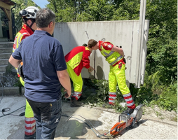 Drei Einsatzkräfte in auffälliger Arbeitskleidung untersuchen ein geschlossenes Gehäuse. Einer der Helfer hat eine Motorsäge in der Nähe. Ein Mann im Vordergrund beobachtet die Situation. Die Umgebung ist grün und es gibt wenig sichtbare Infrastruktur.