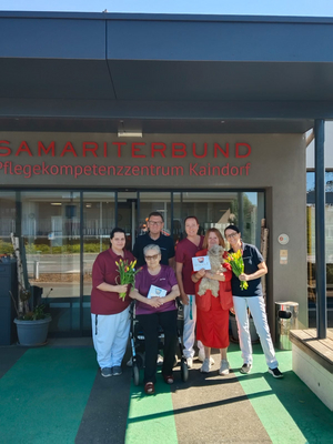 Gruppe von sechs Personen vor dem Eingang des Pflegekompetenzzentrums Kaindorf. Sie halten Blumen und persönliche Karten. Das Schild 