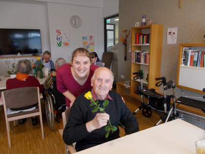 Eine lächelnde Frau umarmt einen älteren Mann mit einer Rose in der Hand.