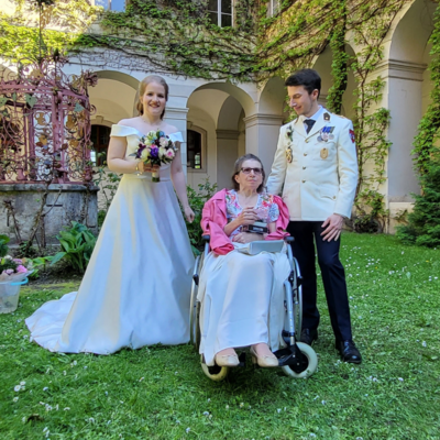 Braut in weißem Kleid steht neben einer Frau im Rollstuhl und einem Mann.