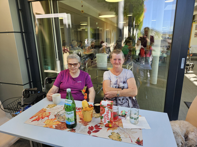 Zwei Frauen sitzen an einem Tisch mit Getränken und Snacks in einem Garten.