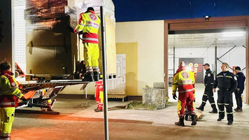 Nachtaufnahme, zwei Sanitäter in Uniform von hinten fotografiert, einer lädt gerade etwas in einen LKW ein, der andere geht zu einer weiteren Gruppe von Menschen 