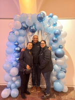 Drei Personen stehen vor einem dekorierten Hintergrund mit blauen Luftballons.