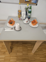 Ein Tisch mit einer hellen Tischdecke, auf dem zwei orangene Schalen stehen, gefüllt mit Leckereien. Darüber hinaus befinden sich zwei Tassen, ein großes Glas und eine Flasche Sprudelwasser. Im Hintergrund ist eine Wand mit einer Pinnwand zu sehen.