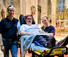 Drei Personen stehen gemeinsam in einer Kirche, eine Person auf einer Trage sitzt.