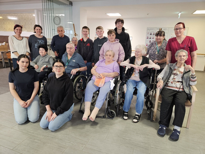 Eine Gruppe von zwölf Personen in einem Gemeinschaftsraum. Ältere Menschen sitzen in Rollstühlen oder auf Stühlen, umgeben von mehreren jungen Menschen. Alle lächeln und stehen in einer freundlichen Pose. Der Raum ist hell und einladend.