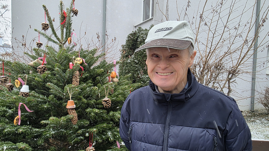 Ein älterer Mann steht lächelnd neben einem dekorierten Weihnachtsbaum.
