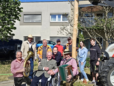 Eine fröhliche Gruppe von Menschen, darunter ein älterer Herr im Rollstuhl, posiert vor einem Gebäude. Einige Personen halten Musikinstrumente wie ein Tuba und ein Akkordeon. Sie stehen in einem Gartenbereich mit ein paar Bäumen und einem Traktor.