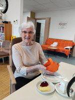 Eine ältere Frau mit kurzen, grauen Haaren sitzt lächelnd an einem Tisch. Sie hält eine orangefarbene Papierbastelei in der Hand. Auf dem Tisch befinden sich Teller mit Marmelade und Butter sowie eine Teekanne. Im Hintergrund ist ein Sofa und ein Wandbild sichtbar.