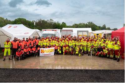 Eine große Gruppe von Einsatzkräften in orange und gelben Uniformen steht vor einem Lager mit Zelten und Fahrzeugen. Sie halten ein Banner mit der Aufschrift 