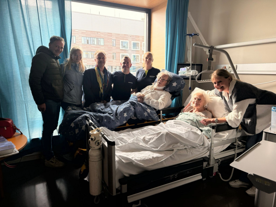 Eine Gruppe von sieben Personen steht in einem Krankenhauszimmer, um eine liegende Patientin im Bett zu besuchen. Die Patientin wird von einer weiteren Person gehalten, beide wirken freundlich. Im Hintergrund sind Fenster und medizinische Geräte sichtbar.
