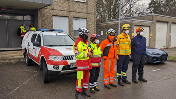 zwei Personen in gelb, roter Samariterbund Uniform, eine PErso in orangener Uniform, zwei weitere Personen ebenso in Katastrophenhilfsdienst Uniform, alle tragen Maske und Schutzhelm inkl. Schutzgläser, dahinter ein Auto der Samariterbund Katastrophenhilfe, zwei weitere Personen in Samariterbund Uniform und Helm die gerade bei einem Gebäude stehen