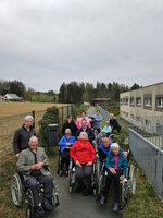 Gruppe von Senioren in Rollstühlen auf einem Gehweg, umgeben von Natur. Einige Personen sitzen, andere stehen hinter ihnen. Ein Gebäude und ein Zaun sind im Hintergrund sichtbar. Es ist bewölkt und die Szene strahlt eine freundliche, gemeinschaftliche Atmosphäre aus.