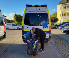 Eine Person küsst einen anderen Menschen im Rollstuhl vor einem Transportfahrzeug.