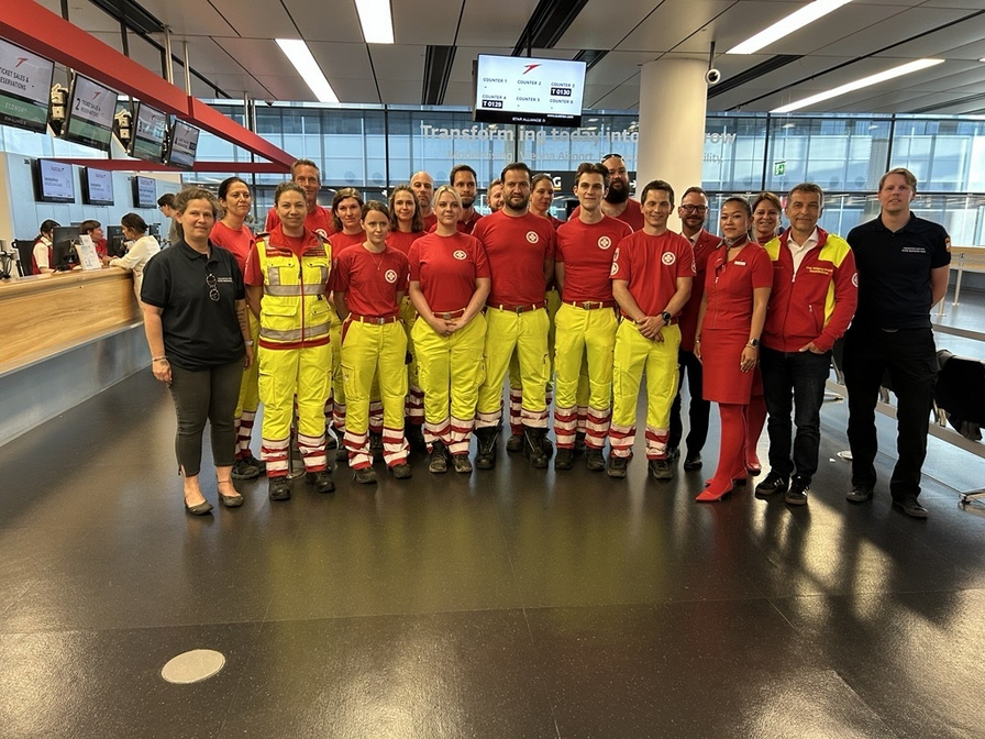 Gruppe von Rettungsdienstmitarbeitern und Mitarbeitern in Uniform, die in einer modernen Empfangshalle stehen. Die Personen lächeln und posieren gemeinsam für ein Foto. Im Hintergrund sind Computerterminals und ein transparenter Bereich mit Fenstern zu sehen.