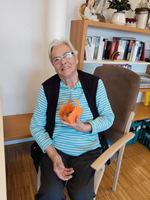 Eine ältere Frau mit grauen Haaren und Brille sitzt in einem Stuhl und hält eine orangefarbene Bastelarbeit in der Hand. Sie trägt ein gestreiftes Oberteil und lächelt freundlich in die Kamera. Im Hintergrund sind Bücherregale und dekorative Elemente zu sehen.