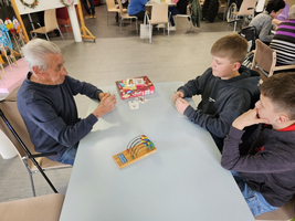 Ein älterer Mann spielt mit zwei Kindern an einem Tisch. Auf dem Tisch liegen Spielkarten und ein Beschäftigungsspiel. Im Hintergrund sind mehrere Personen zu sehen, die ebenfalls beschäftigt sind. Die Atmosphäre wirkt freundlich und einladend.