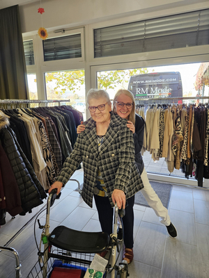 Eine ältere Frau mit Rollator steht lächelnd vor einer Kleiderboutique, umarmt von einer jüngeren Frau.