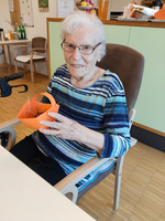 Eine ältere Frau mit Brille sitzt auf einem Stuhl und hält einen orangefarbenen Korb in der Hand. Sie trägt ein gestreiftes Oberteil und lächelt freundlich in die Kamera. Im Hintergrund sind Tische und Getränke sichtbar.