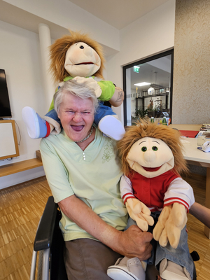 Eine lächelnde Frau sitzt mit zwei Puppen auf ihrem Schoß.